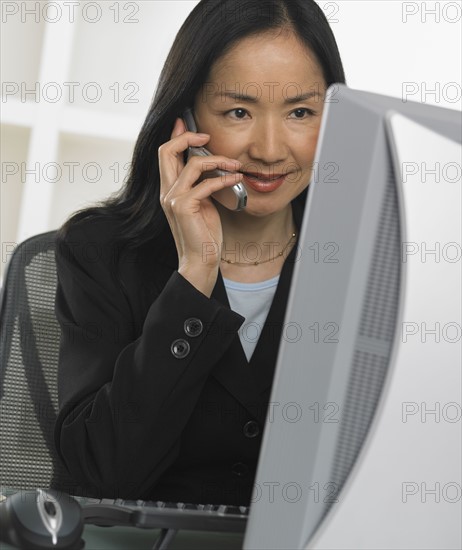 Asian businesswoman using cell phone at computer.