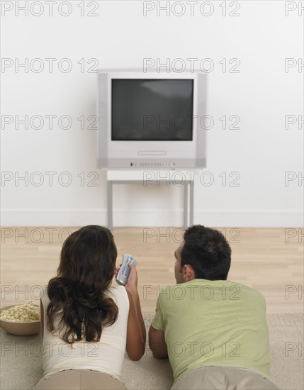 Couple laying on floor with popcorn watching television .