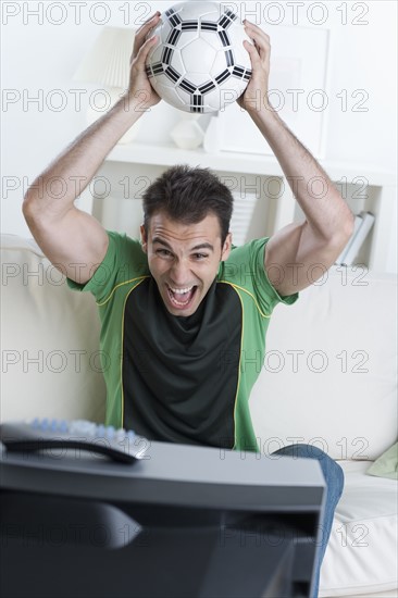 Man cheering and holding soccer ball while watching television.