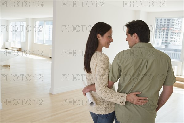 Couple hugging and holding blueprints in empty house.