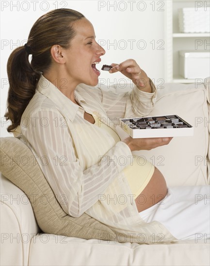 Pregnant woman on sofa eating box of chocolates.