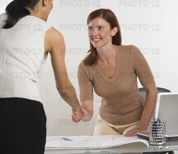 Two businesswomen shaking hands over blueprints.