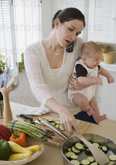 Mother holding baby, talking on telephone and cooking.