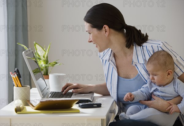 Mother typing on laptop and holding baby.