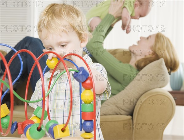 Young son playing with block toy with mother and baby in background. Date : 2006