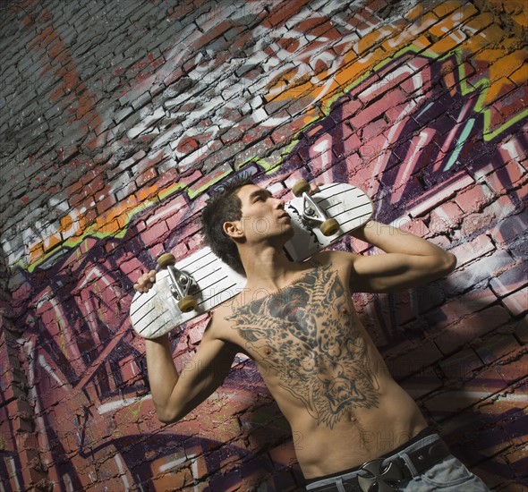 Asian man holding skate board in front of graffitied wall. Date : 2007