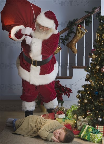 Santa Claus looking at boy sleeping next to Christmas tree.