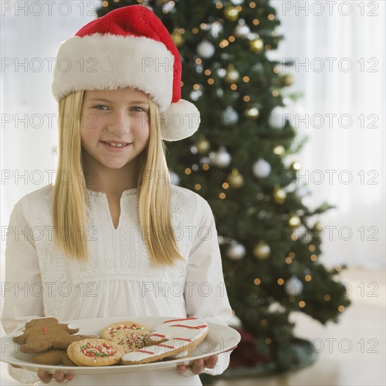 Girl in Santa Claus hat holding plate of cookies.