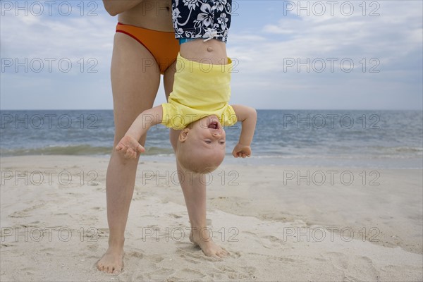 Mother holding baby boy upside-down on beach. Date : 2008