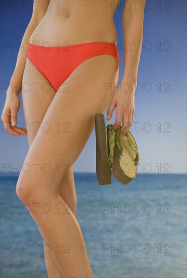 Midsection of woman in bikini holding flip-flops on beach.