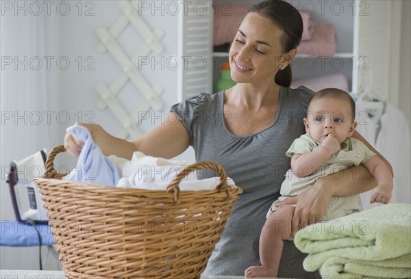 Mother holding baby son and sorting laundry.