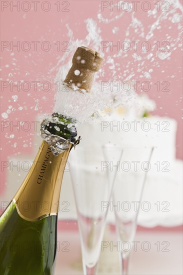 Cork exploding from champagne bottle with wedding cake in background.