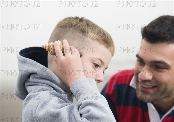 Father watching son hold seashell up to ear. Date : 2008