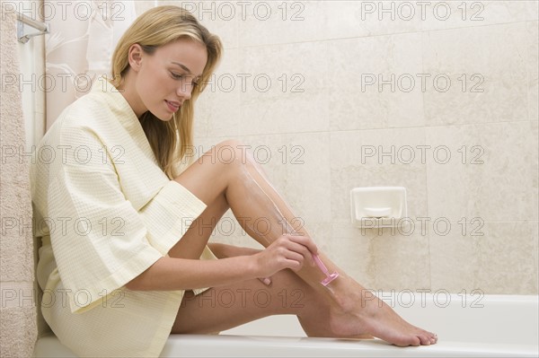 Woman shaving legs on edge of bathtub.