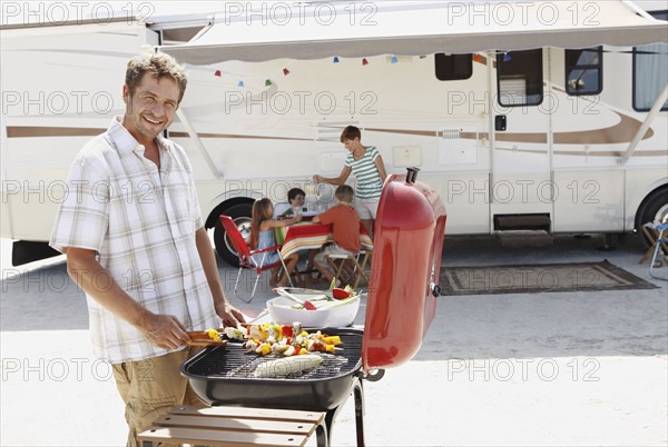 Man barbecuing with family and motor home in background. Date: 2008