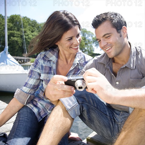 Couple sitting on dock looking at photos on digital camera. Date: 2008