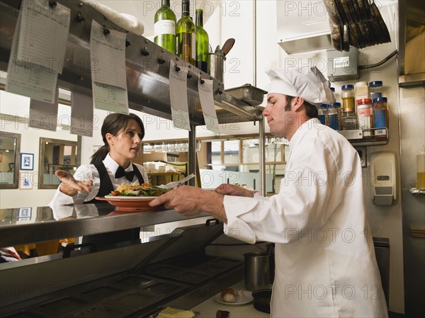 Waitress gesturing to chef in restaurant kitchen.