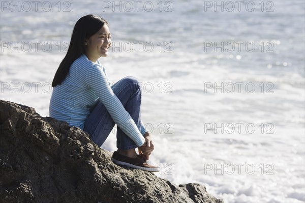 Teenage girl sitting on coastal rock. Photographe : PT Images