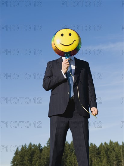 Businessman holding smiley face balloon over face.