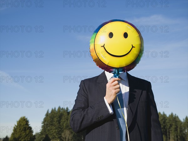 Businessman holding smiley face balloon over face.