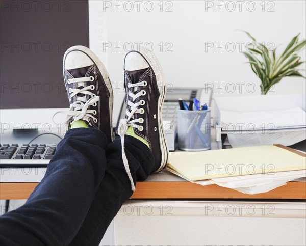Young office worker in casual clothes relaxing in office with legs on desk. Photographe : Jamie Grill