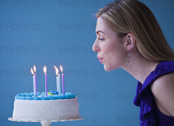 Young woman blowing candles on birthday cake, studio shot. Photographe : Daniel Grill