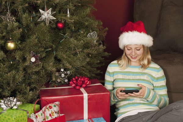 Teenage girl (13-15) sitting in front of Christmas tree and text messaging. Photographe : Sarah M. Golonka