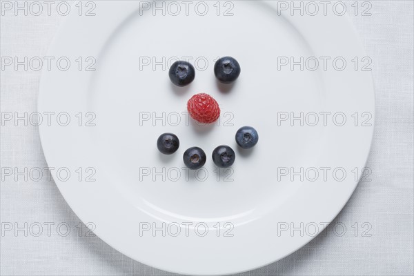 Smiley face of blueberries and raspberry on white plate. Photographe : Kristin Lee