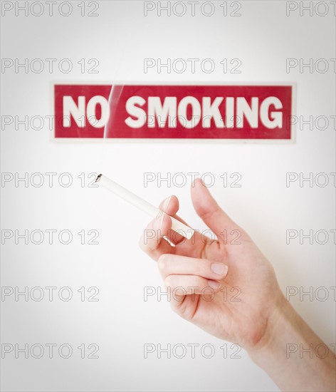 Hand holding cigarette in front of no smoking sign. Photographe : Jamie Grill