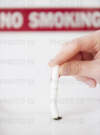 Hand putting out cigarette in front of no smoking sign. Photographe : Jamie Grill