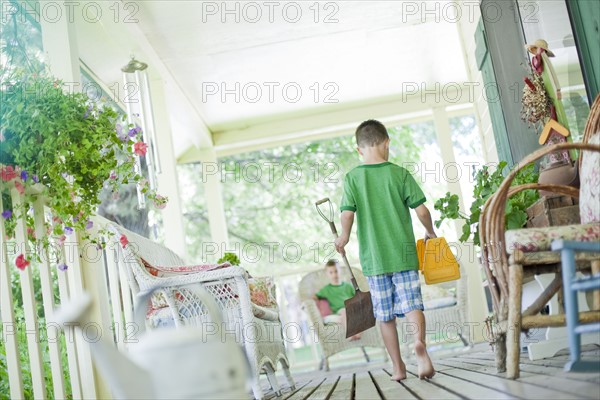 Young boy carrying shovel and tool box on porch. Photo. Tim Pannell