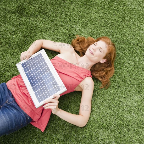 Beautiful woman lying on grass while holding solar panel. Photo : Jamie Grill