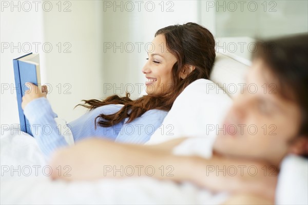 Woman reading book in bed beside sleeping husband. Photo. momentimages