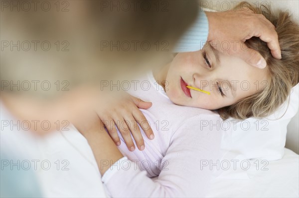 Grandfather tending granddaughter (10-11) lying in bed with thermometer in mouth. Photo : Momentimages