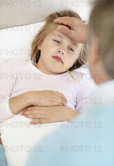 Grandfather tending granddaughter (10-11) lying in bed with thermometer in mouth. Photo : Momentimages