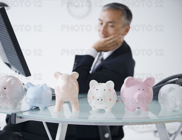 Businessman looking at piggybanks standing on desk. Photo : Jamie Grill