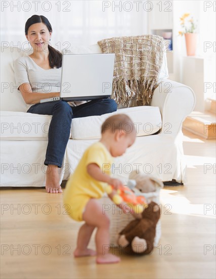 Baby boy (6-11 months) playing, mother sitting on sofa and using laptop. Photo : Daniel Grill