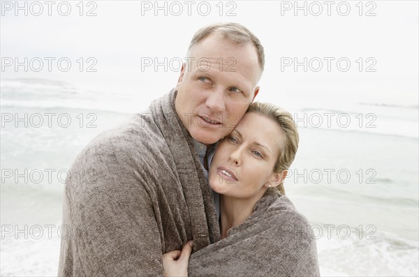 Embraced couple covered by blanket looking away on beach. Photo : Momentimages