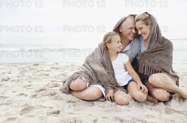 Smiling parents with daughter (10-11) on coastline covered by blanket. Photo : Momentimages