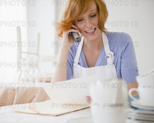 Young woman in apron talking on phone in kitchen. Photo : Jamie Grill