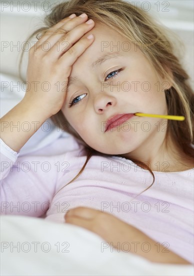 Sad girl (10-11) lying in bed with thermometer in mouth. Photo : Momentimages