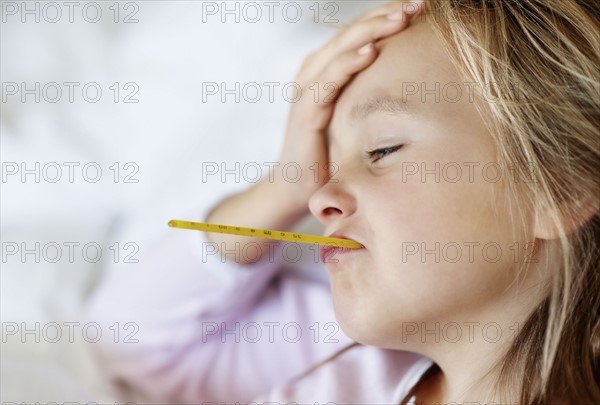 Sad girl (10-11) lying in bed with thermometer in mouth. Photo : Momentimages