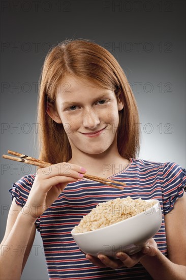 Portrait of redhead girl (10-11) holding chopsticks and bowl of rice, studio shot. Photo : FBP