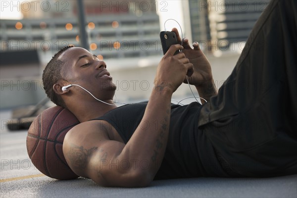 USA, Utah, Salt Lake City, Young man lying on basketball and listening to mp3 player. Photo : Mike Kemp