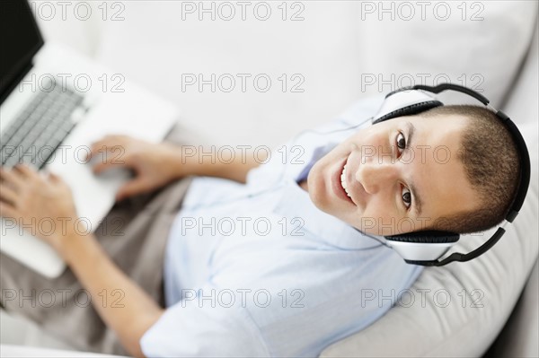 South Africa, Young man wearing headphones sitting on sofa and using laptop, high angle view. Photo : momentimages