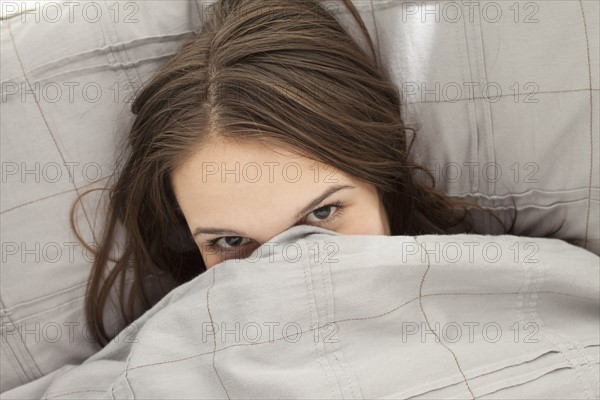 Portrait of young woman lying in bed hiding under bedding. Photo : Mike Kemp