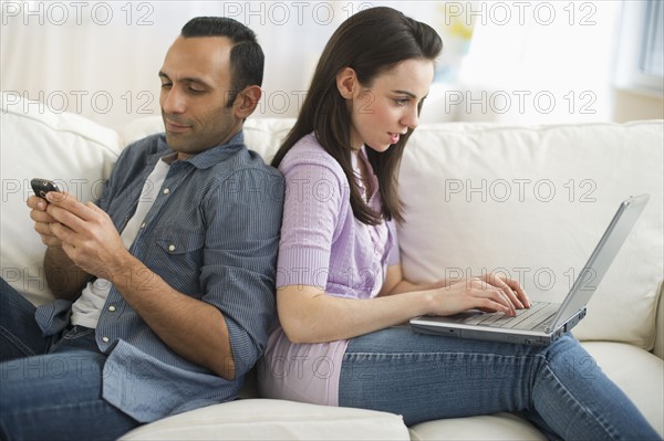 Couple sitting on sofa back to back using laptop and phone.