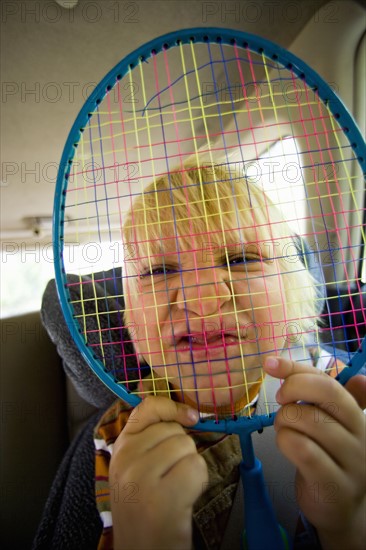 Portrait of boy (6-7) holding badminton racquet over face. Photo : Noah Clayton