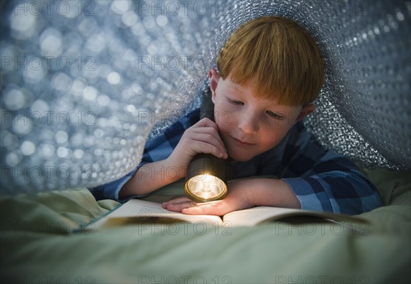 Boy (8-9) reading book under bed covers. Photo : Jamie Grill Photography