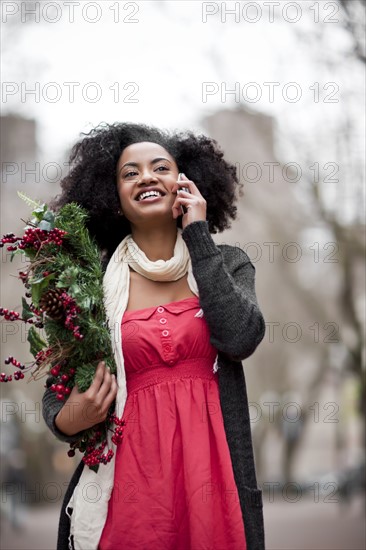 USA, Washington State, Seattle, Cheerful young woman holding christmas wreath while using mobile phone. Photo : Take A Pix Media